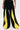 A person wears the GAME TIME DECISION DENIM PANT—wide-leg black jeans with bold yellow panels down the front and sides, featuring a drawstring waist. Hands rest at their sides against a plain, light background.