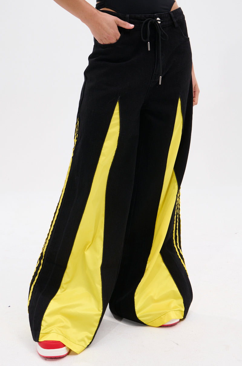 Wearing the GAME TIME DECISION DENIM PANT with bold yellow inserts and red-and-white sneakers, a person stands against a plain white background with one hand in their pocket.