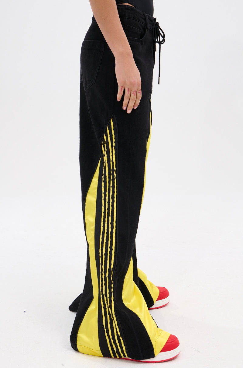 A person models the GAME TIME DECISION DENIM PANT—black wide-leg jeans with bold yellow panels and three vertical yellow stripes—paired with red and white sneakers, shown from the waist down against a plain white background.