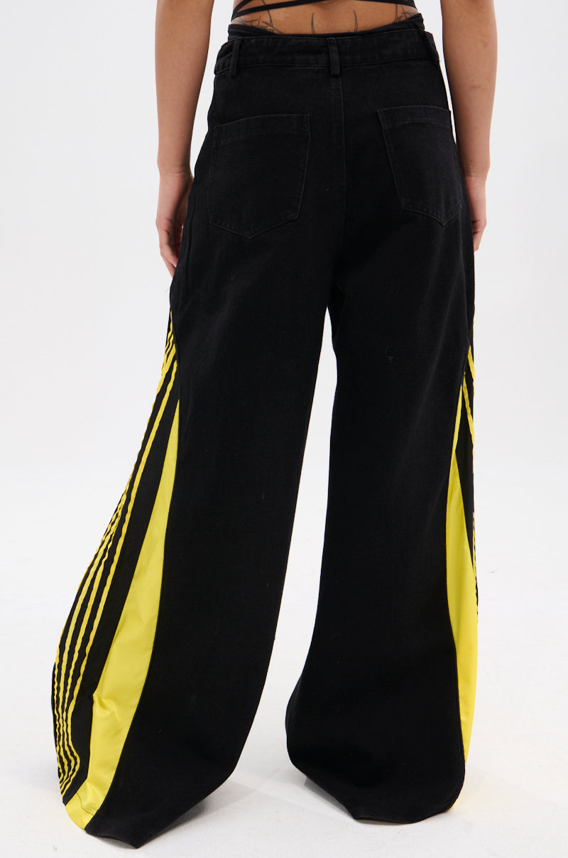 A person is seen from the back wearing GAME TIME DECISION DENIM PANT—black wide-leg streetwear jeans featuring bright yellow panels and black side stripes—against a plain white background.