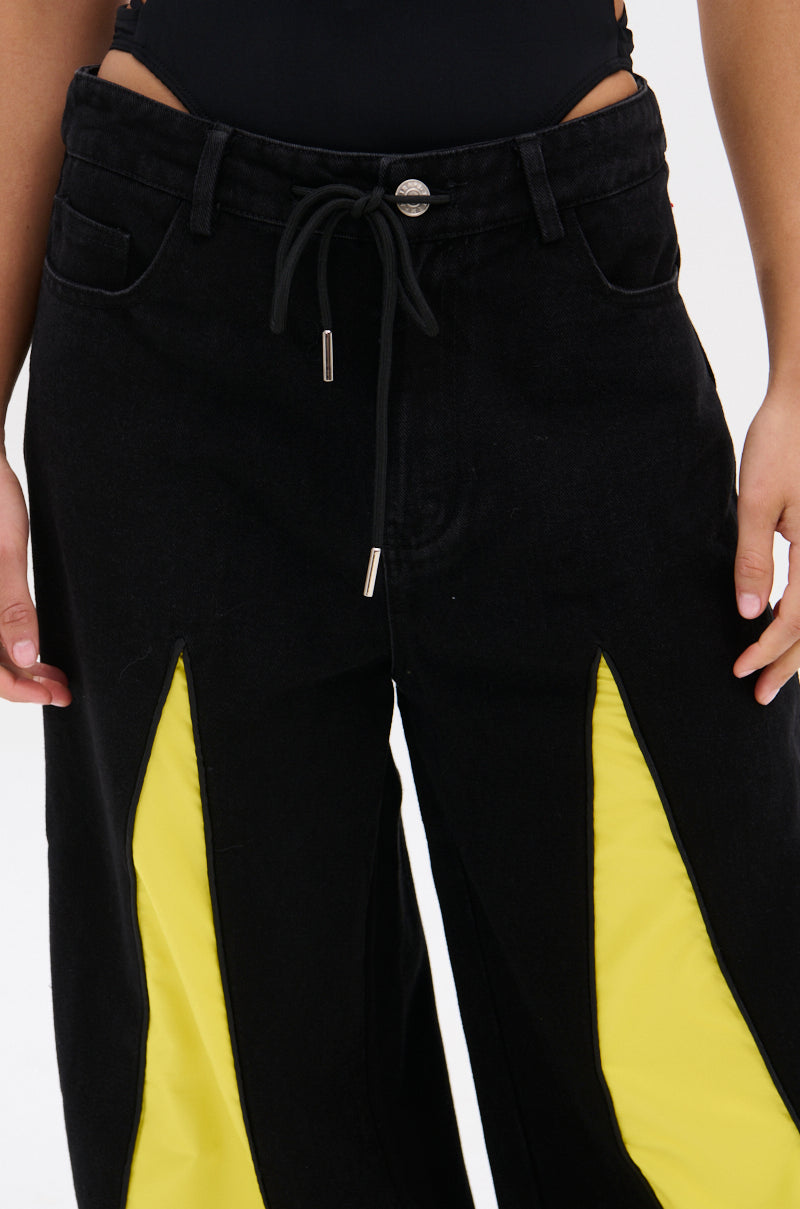 A person modeling the GAME TIME DECISION DENIM PANT—black streetwear jeans with a drawstring waist and bold yellow triangular insets on the front thighs—shown from waist to mid-thigh, hands relaxed at their sides.