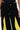 A person modeling the GAME TIME DECISION DENIM PANT—black streetwear jeans with a drawstring waist and bold yellow triangular insets on the front thighs—shown from waist to mid-thigh, hands relaxed at their sides.