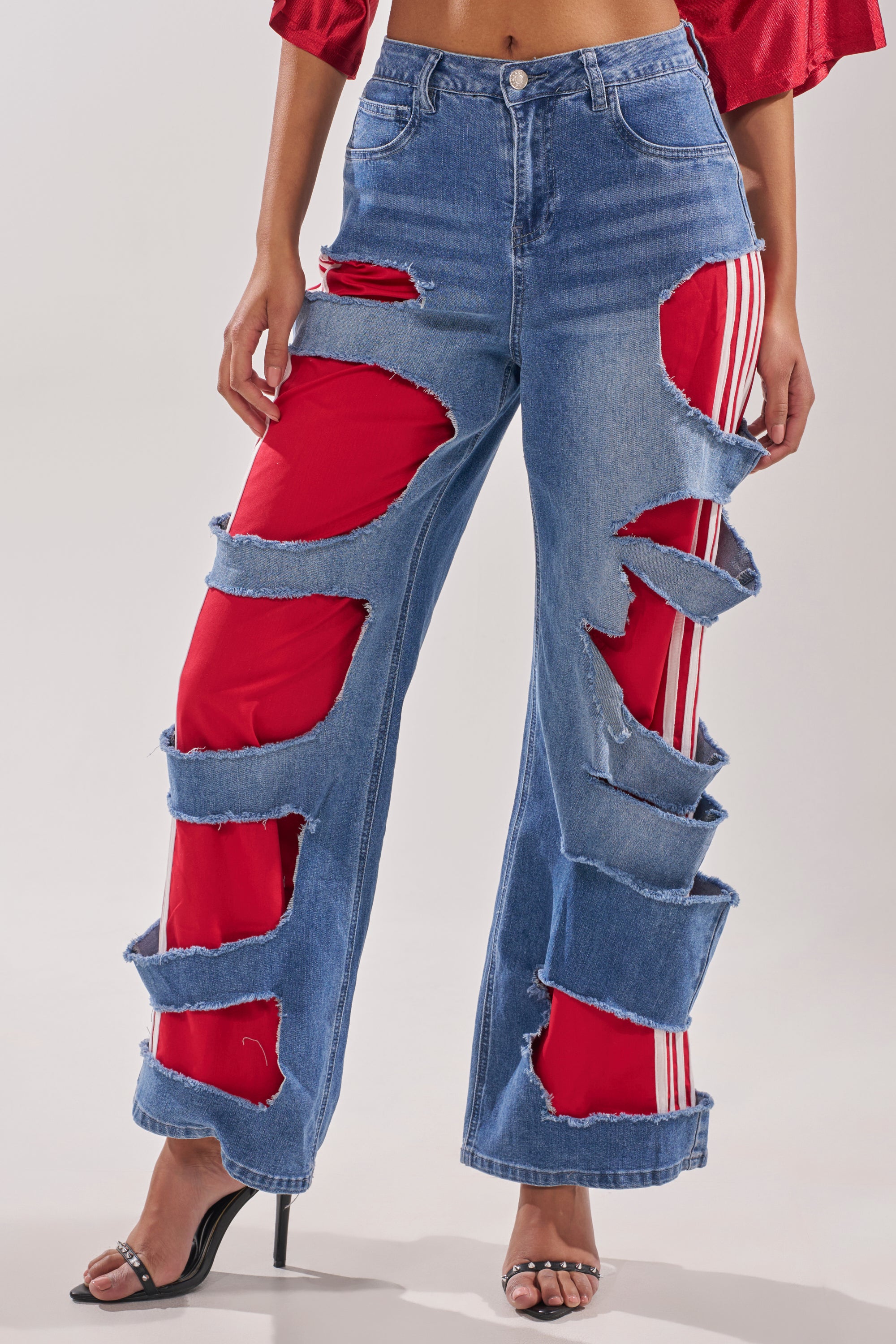 A person wears the LEXIE LOW RISE DENIM TRACK PANT—distressed blue jeans with white-striped track pants beneath—styled with black high-heeled sandals and a red cropped top. 0