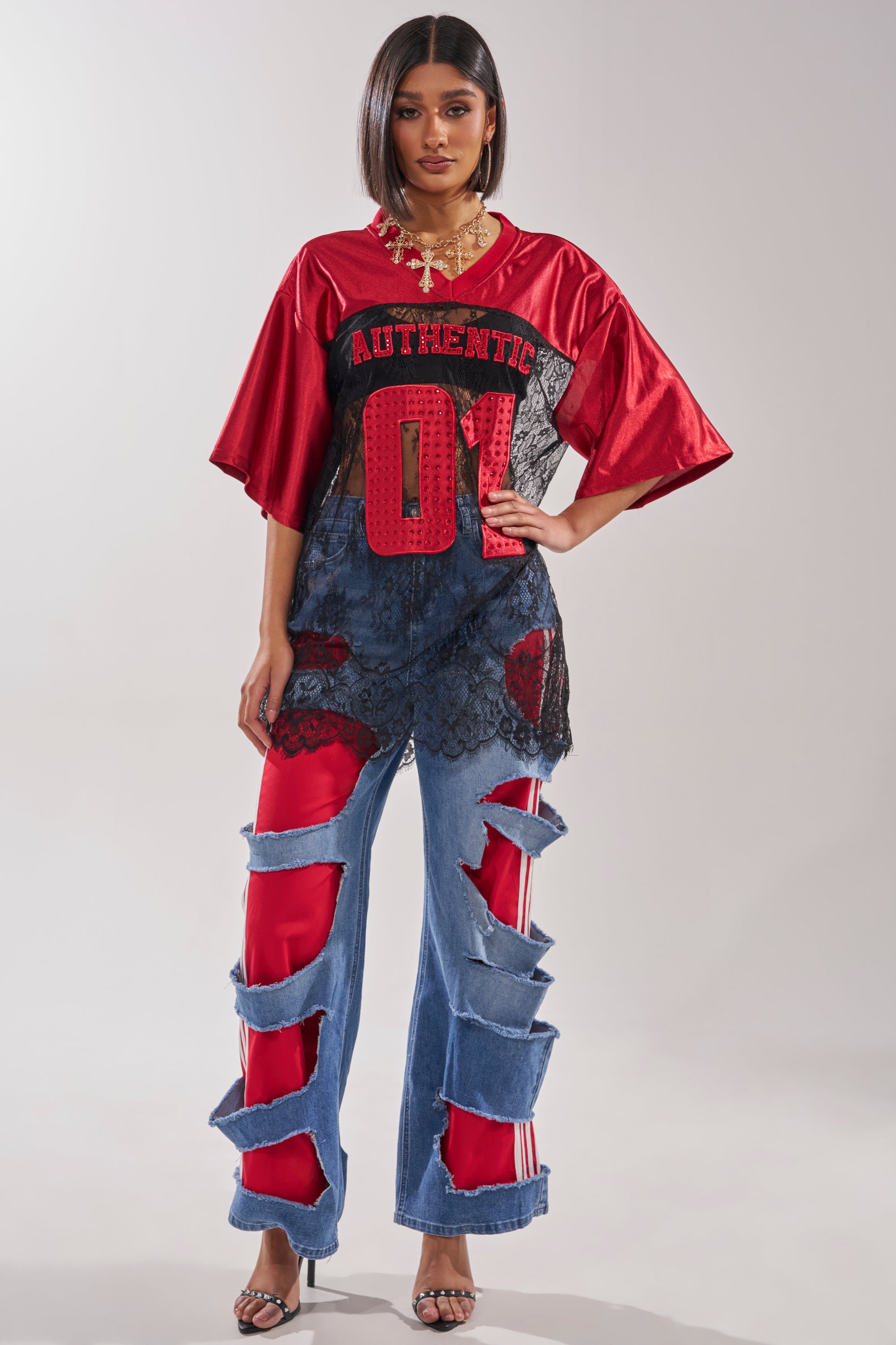 A woman stands confidently in a red “AUTHENTIC 01” jersey, black heels, and LEXIE LOW RISE DENIM TRACK PANT—Y2K-inspired ripped blue jeans lined with red fabric. She wears layered necklaces and has straight hair. 1