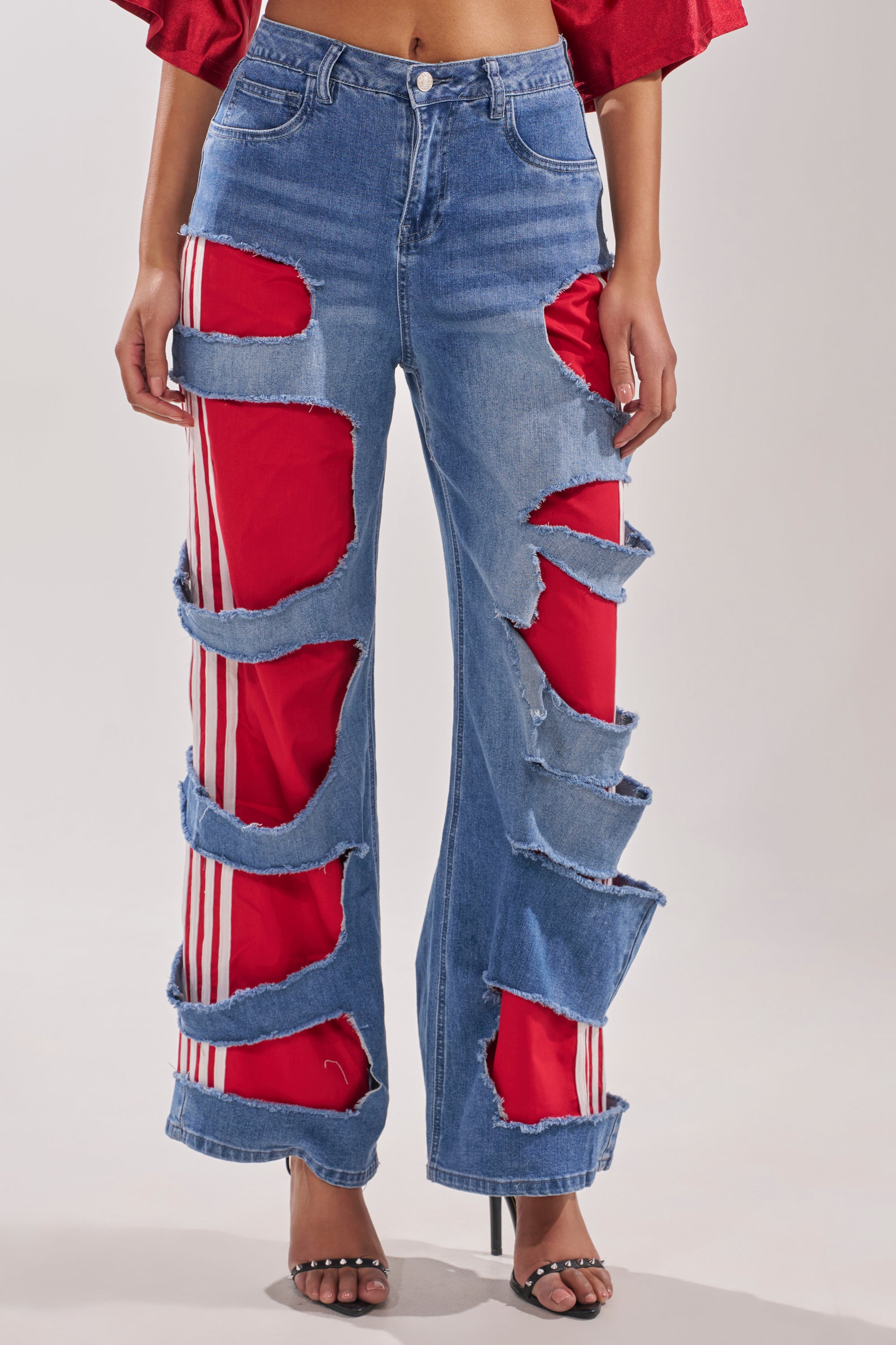 A person wears the LEXIE LOW RISE DENIM TRACK PANT with large, irregular rips showing red-and-white striped fabric beneath, styled with a red top and black open-toe heels. 2