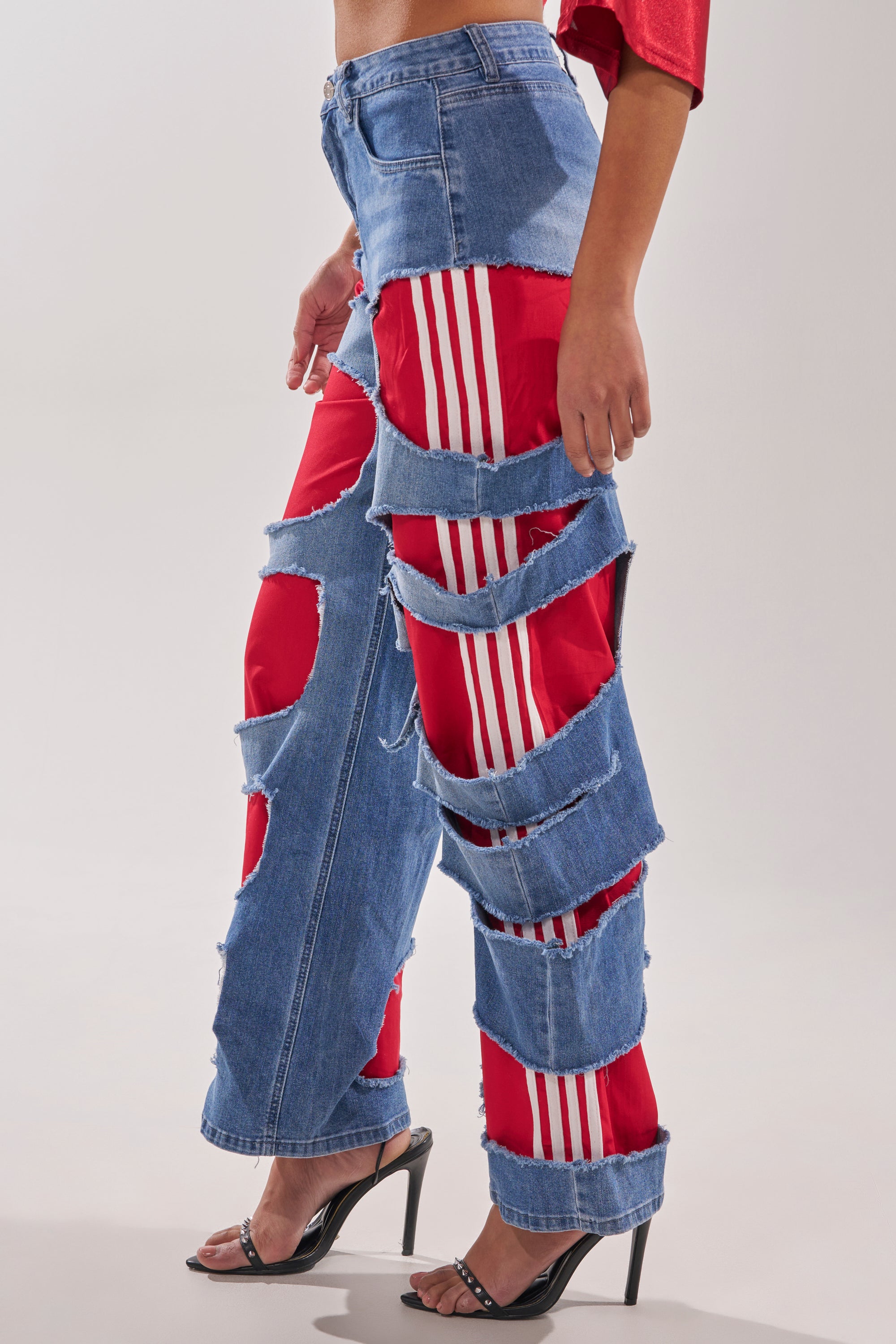 Wearing LEXIE LOW RISE DENIM TRACK PANT featuring blue denim with bold horizontal rips that reveal red Y2K-style track pants with white stripes underneath, paired with black heeled sandals. 3