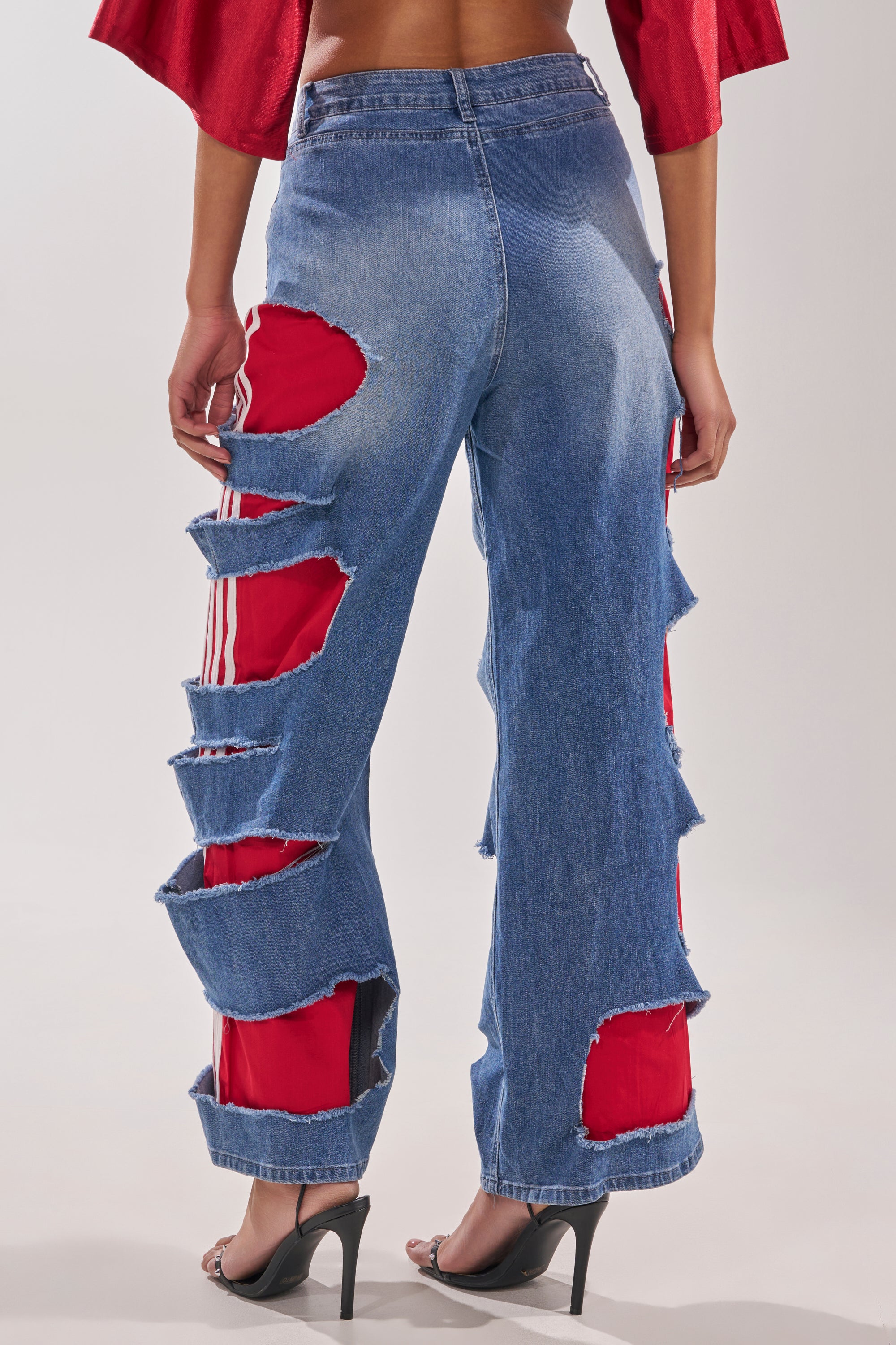 A person wearing the LEXIE LOW RISE DENIM TRACK PANT, featuring wide-leg blue jeans with large horizontal rips that reveal Y2K-style track pants beneath, paired with black high-heeled sandals. Photo taken from behind. 4