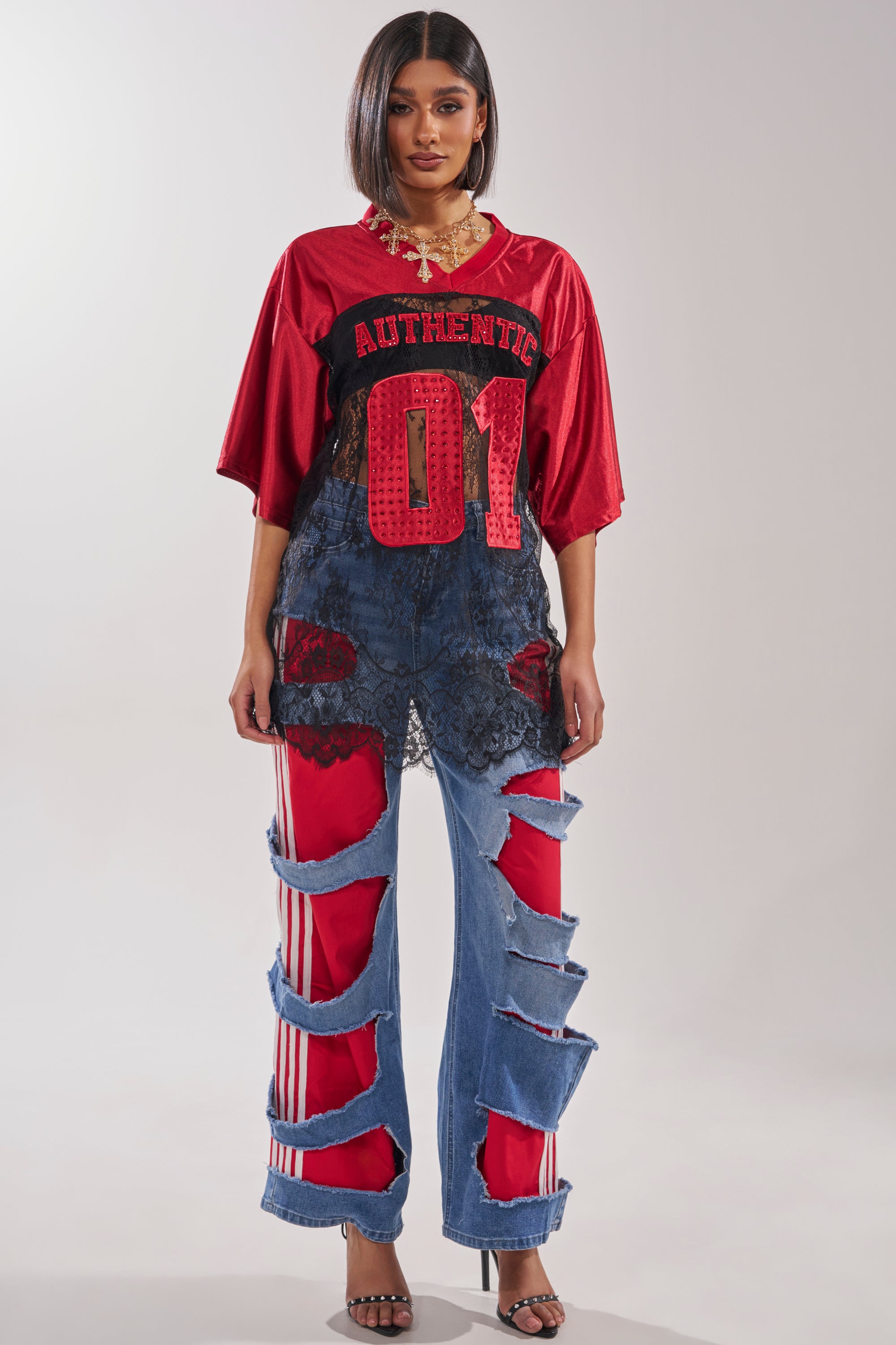 A woman wears a loose red sports jersey with "AUTHENTIC 01" over a lace top, paired with the LEXIE LOW RISE DENIM TRACK PANT featuring large ripped details with red fabric beneath, and black heeled sandals. 5