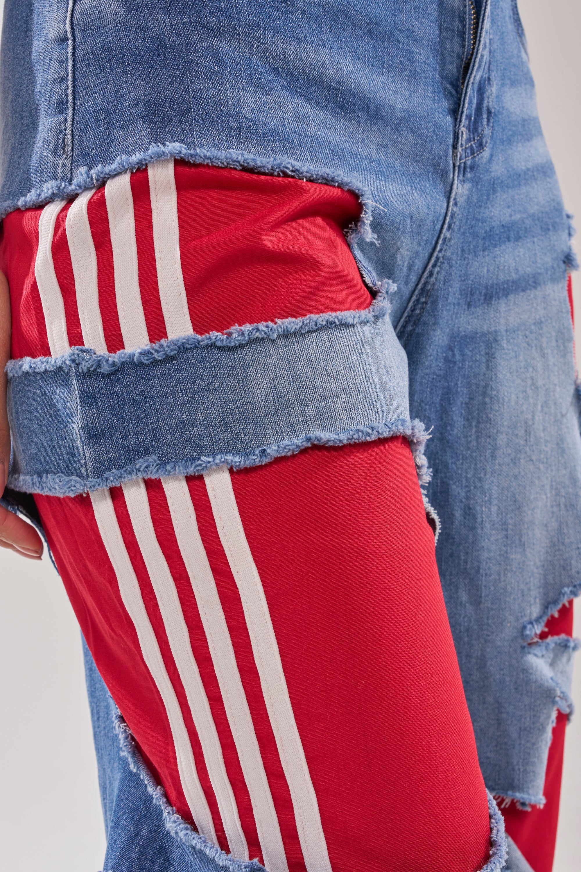A close-up of a leg wearing the LEXIE LOW RISE DENIM TRACK PANT, featuring blue denim with large rips that reveal red pants with white stripes underneath—a striking Y2K streetwear-inspired look. 8