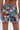 A person wearing the KISS MY PATCH DENIM SHORT, vintage-wash shorts adorned with colorful patches—flowers, cherries, peace signs, an alien, and quirky slogans. The frayed hems and bold designs create a playful, standout look.