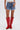 Wearing the KISS MY PATCH DENIM SHORT with colorful patches, a white ribbed tank top, and red knee-high cowboy boots with white embroidery, a person stands against a plain light background.