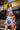 A young woman stands outside a building wearing the KISS MY PATCH DENIM SHORT, featuring colorful patches, and has a rainbow-striped crochet sweater draped over her shoulders as she looks back at the camera.