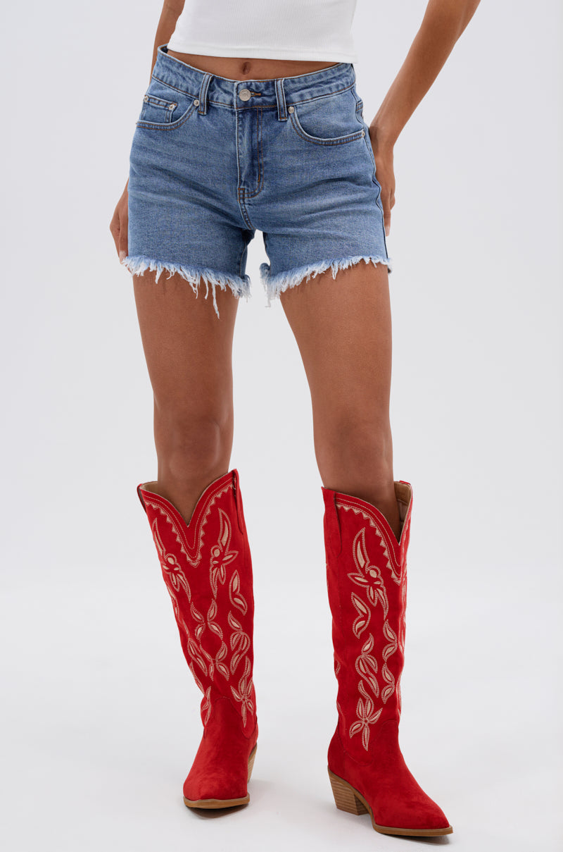 Wearing KISS MY PATCH DENIM SHORT, a white top, and tall red cowboy boots with tan embroidery, a person stands against a plain light background.
