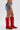 A person stands sideways wearing the KISS MY PATCH DENIM SHORT, featuring colorful patches and embroidery, paired with tall red cowboy boots with decorative stitching. The background is plain white.
