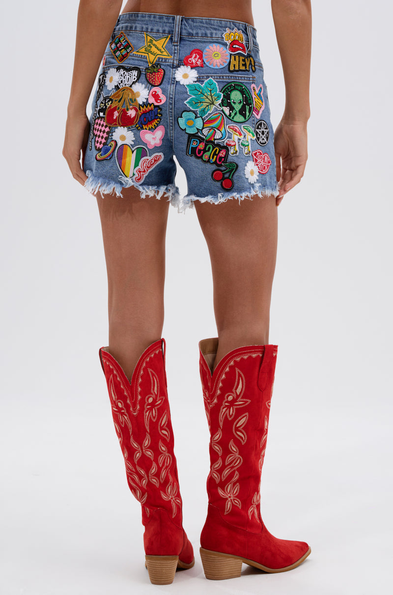 Wearing the KISS MY PATCH DENIM SHORT with vibrant patches and embroidery, paired with tall red cowboy boots with white stitching. Photo is taken from behind against a plain background.