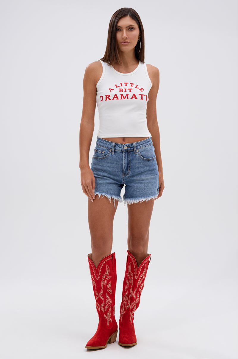A woman with straight brown hair and a neutral expression wears a white tank with red "A LITTLE BIT DRAMATIC" text, red cowboy boots with white embroidery, and the KISS MY PATCH DENIM SHORT.