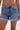 Wearing KISS MY PATCH DENIM SHORT—vintage-wash blue shorts with frayed hems and colorful patches—a person stands against a plain background. The image is cropped to show only the lower torso and upper legs.