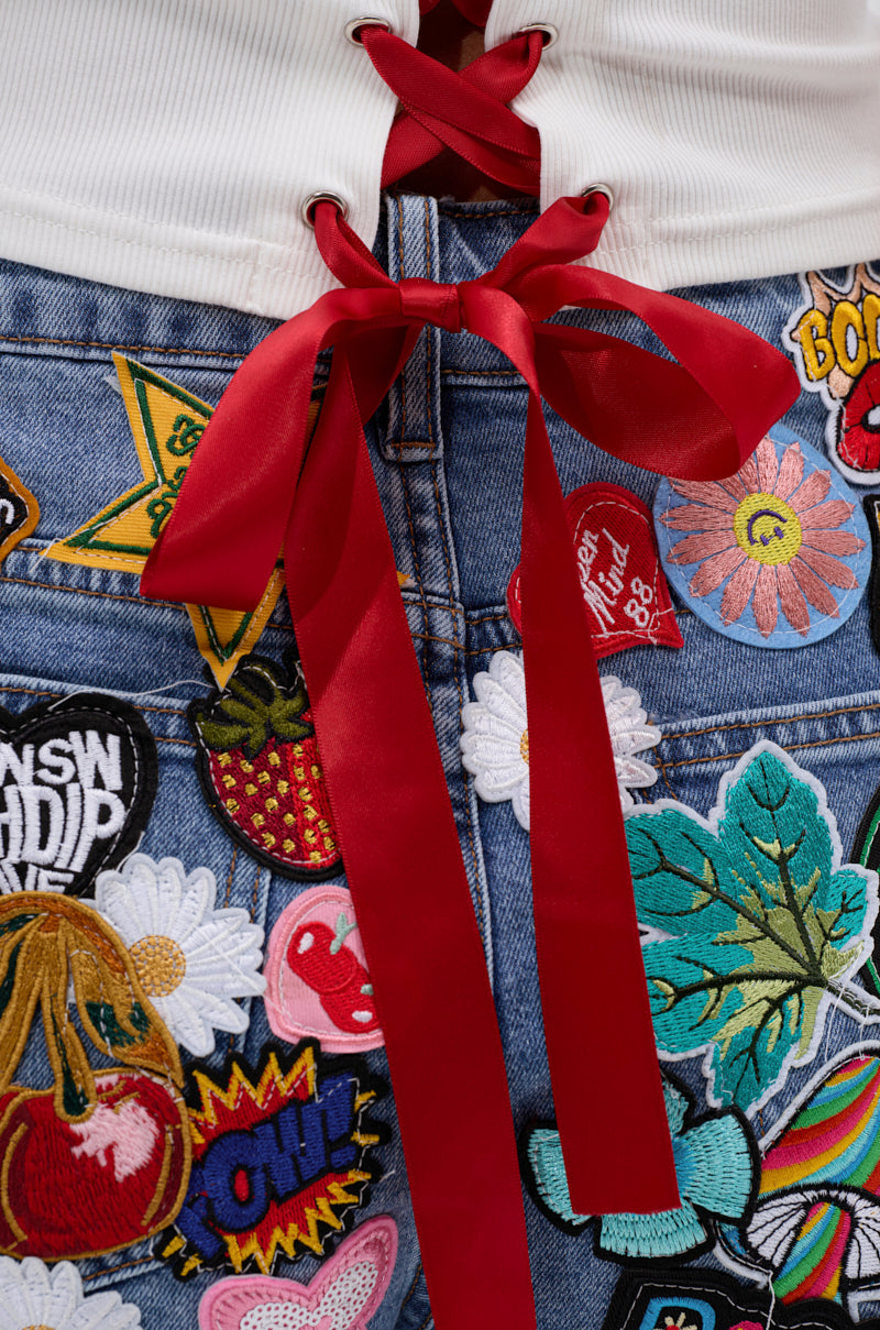 Close-up of the KISS MY PATCH DENIM SHORT, featuring vintage-wash denim with colorful patches—flowers, leaves, fruit, and words. Styled with a white top that has a red ribbon lace-up bow above the shorts.