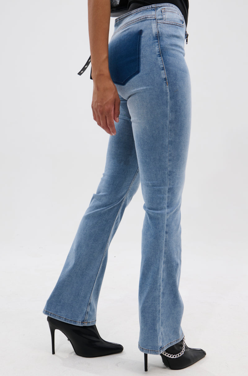 Model wearing the TOO GOOD TO BE TRUE LOW RISE DENIM FLARE PANT in light blue with a dark blue back pocket, paired with black pointed-toe high-heeled boots featuring silver chain detail, on a white background.