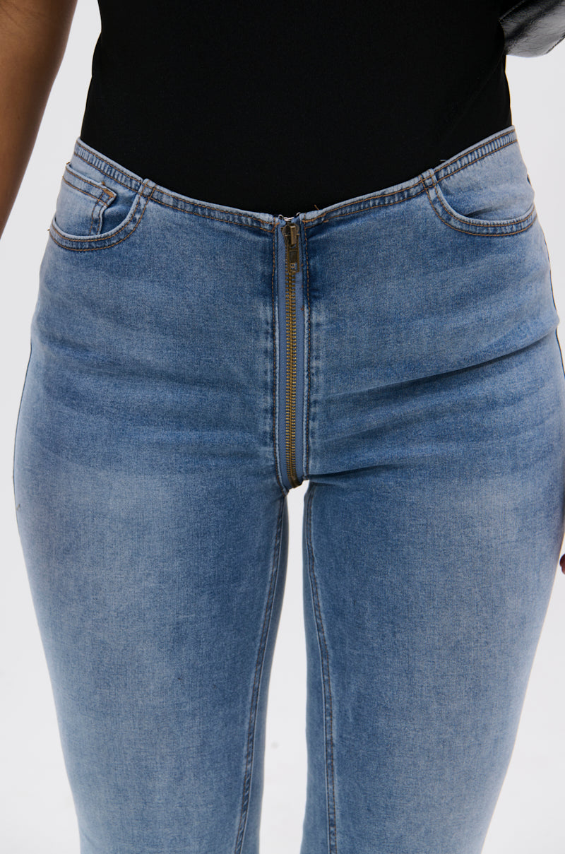 A person models the TOO GOOD TO BE TRUE LOW RISE DENIM FLARE PANT, featuring a visible front zipper, paired with a black top and shown from the waist down against a plain white background.