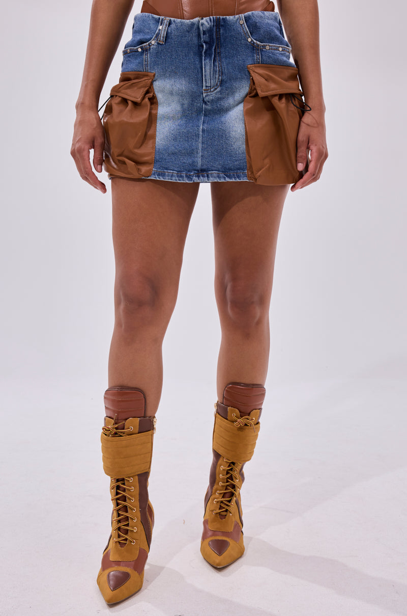 A person in the ROCK MY WORLD DENIM MINI SKIRT, a blue high-rise mini with brown cargo pockets, pairs it with matching brown lace-up boots. Photo cropped from waist to mid-calf, standing against a plain background.