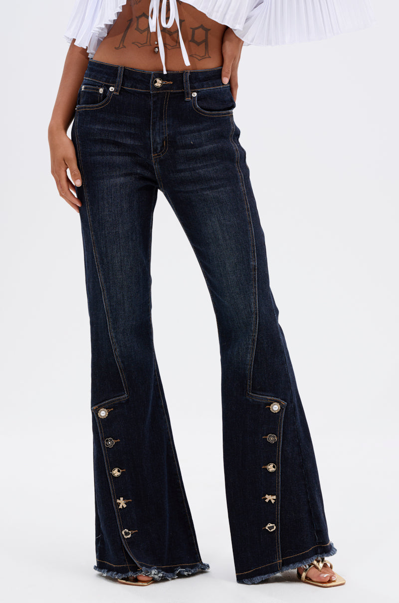 Wearing the AUDREY FLARE DENIM PANT—high-rise, dark wash flare jeans with front decorative buttons and frayed hems—paired with an open white crop top. The person’s face is not visible.