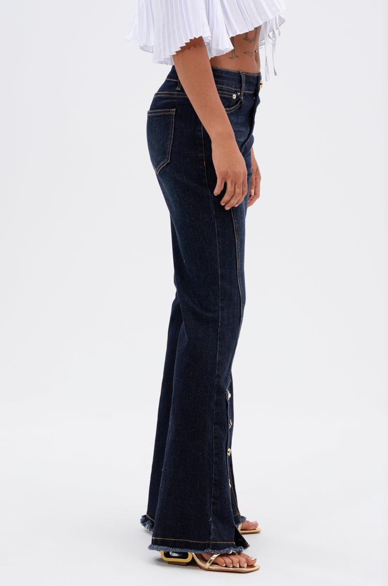 A person stands sideways on a white background, showing their profile from shoulders to feet, wearing the AUDREY FLARE DENIM PANT in high-rise dark wash with side buttons and a white pleated crop top.
