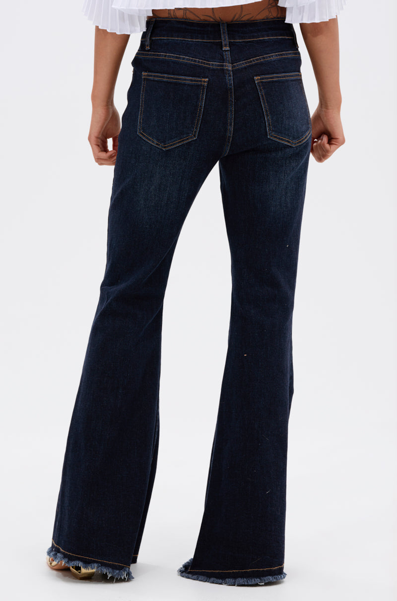 Wearing the AUDREY FLARE DENIM PANT, a high-rise, dark wash style with visible back pockets and raw, frayed hems. Photo is taken from behind, highlighting the jeans from waist to feet.