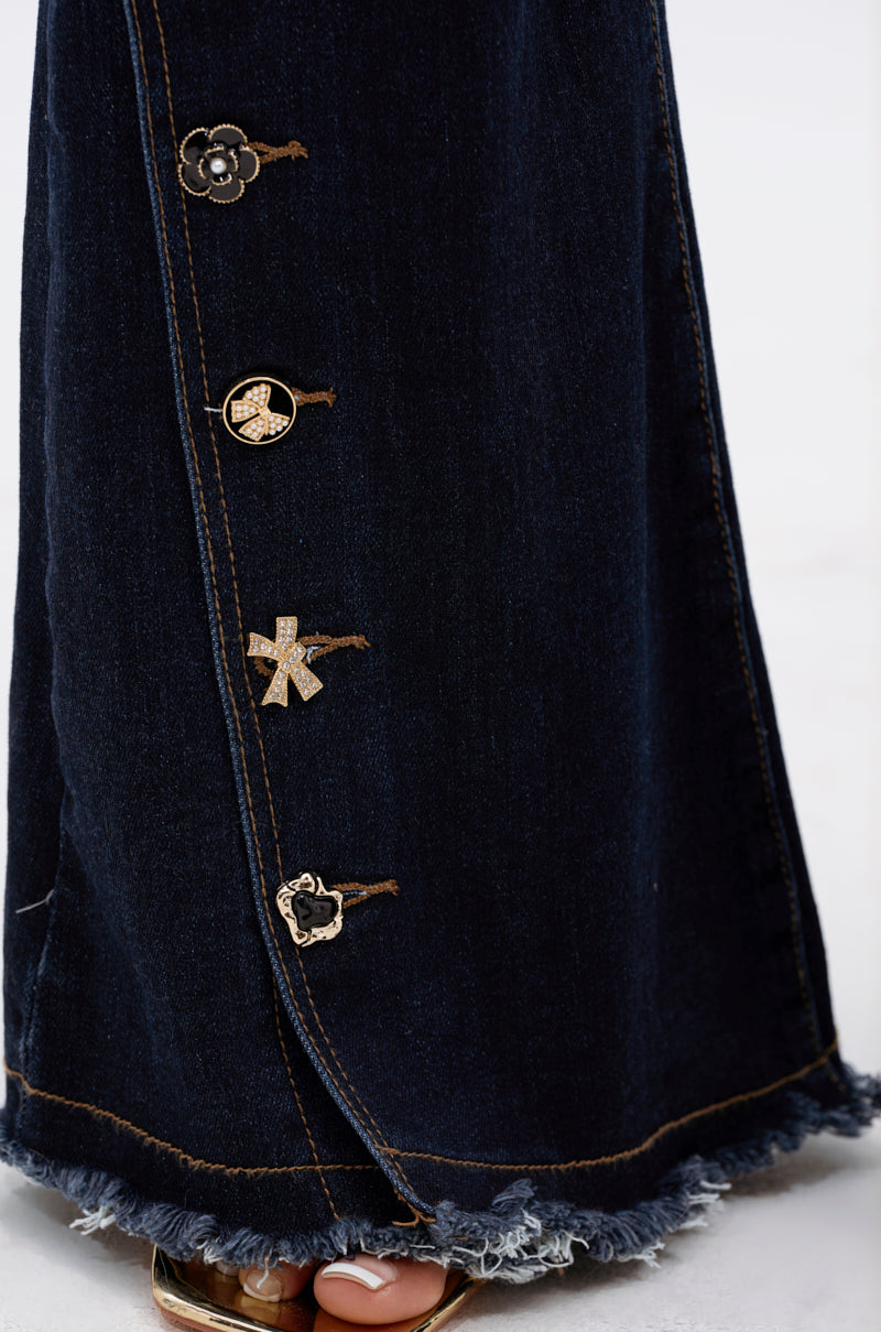 A close-up of the AUDREY FLARE DENIM PANT in dark wash, featuring frayed hems and metallic buttons along the side seam, styled with open-toed sandals.