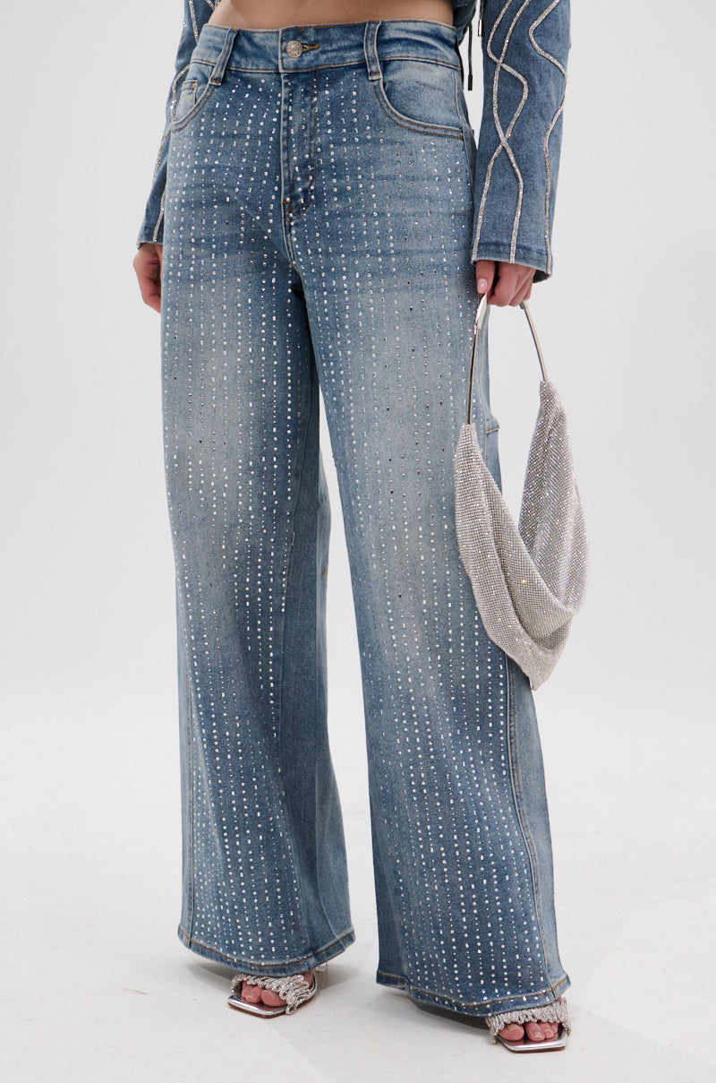 A person wearing the SPARKLE SHINE DENIM PANT—high-rise, wide-leg jeans embellished with vertical rhinestone rows—paired with a matching rhinestone handbag, open-toe heeled sandals, and a long-sleeve top with decorative stitching.