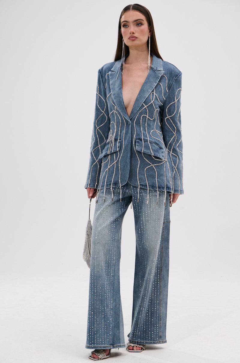 A woman with long, straight brown hair wears SPARKLE SHINE DENIM PANT—a blue, wide-leg style with decorative beaded lines—paired with a matching pantsuit, silver high-heeled sandals, and a small silver handbag against a plain background.