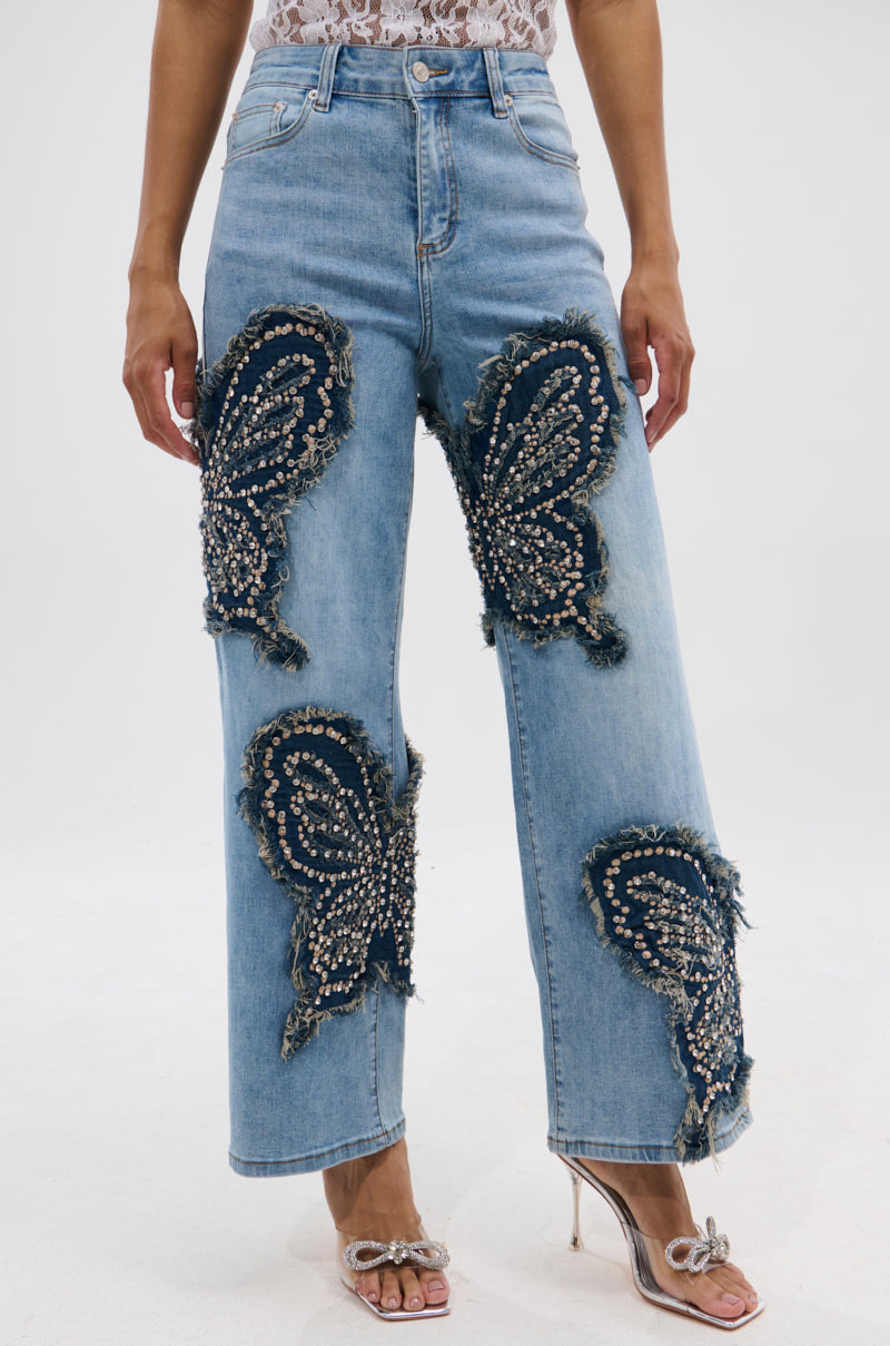 A person wears REBIRTH BUTTERFLY PATCH JEANS in light blue, featuring large beaded and fringed butterfly patches, styled with a lace top and open-toed heeled sandals. Only the lower body is shown. 0