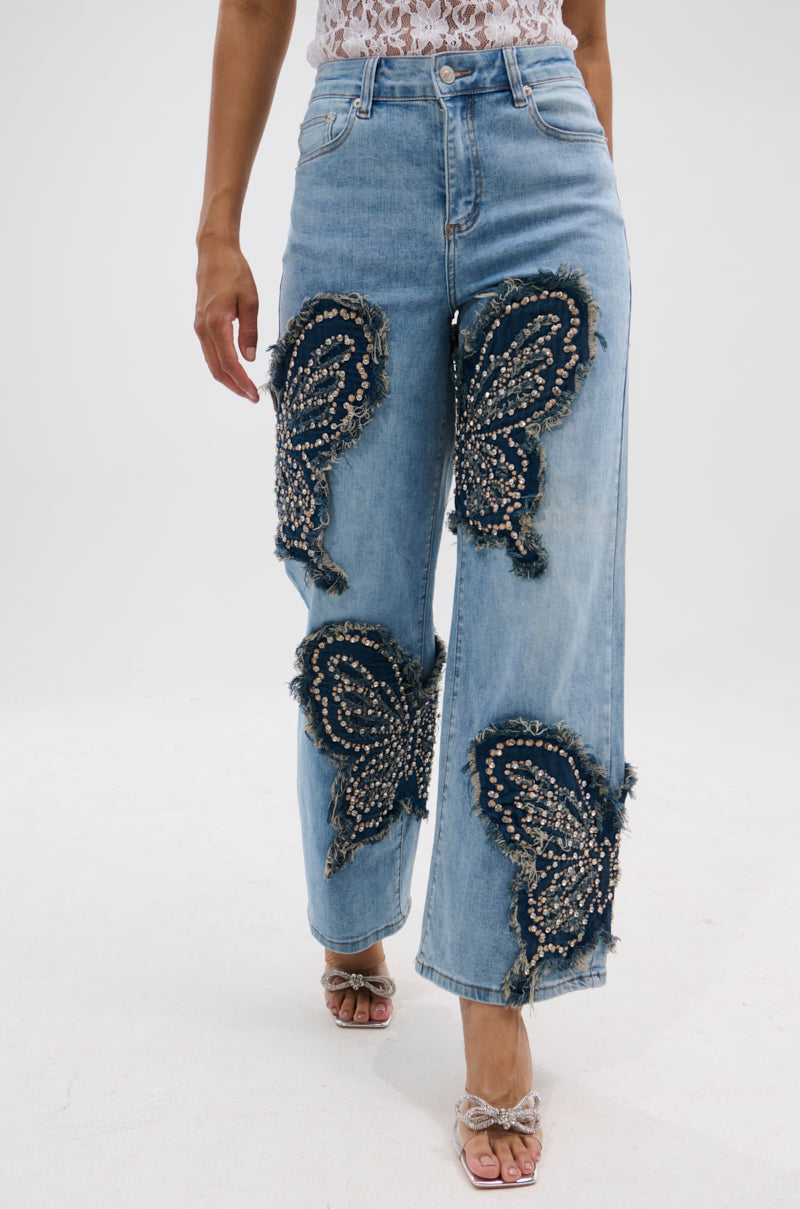 A person wears the REBIRTH BUTTERFLY PATCH JEANS—light blue, high-rise straight-leg jeans featuring large dark paisley studded patches with frayed edges—paired with open-toe heels and a white lace top. 1