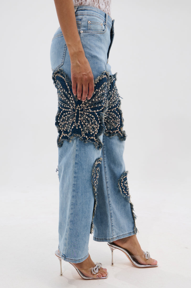 A person wears REBIRTH BUTTERFLY PATCH JEANS featuring large dark denim butterfly appliqués with pearls and rhinestones, paired with silver high-heeled sandals, standing against a white background. 2