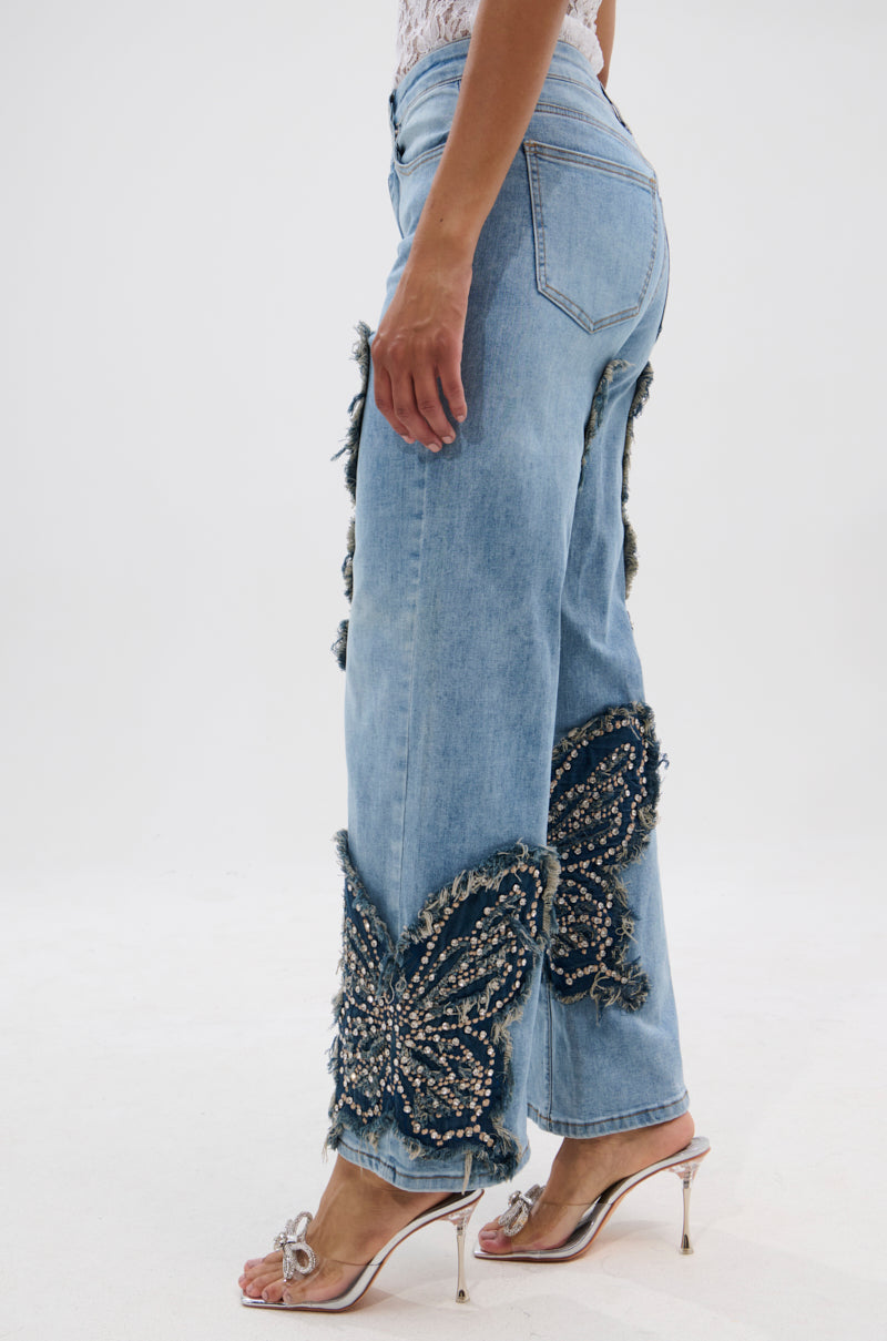 A person wears REBIRTH BUTTERFLY PATCH JEANS with high-rise straight legs, large frayed beaded butterfly patches, paired with clear heeled sandals and a white lace top against a plain white background. 3