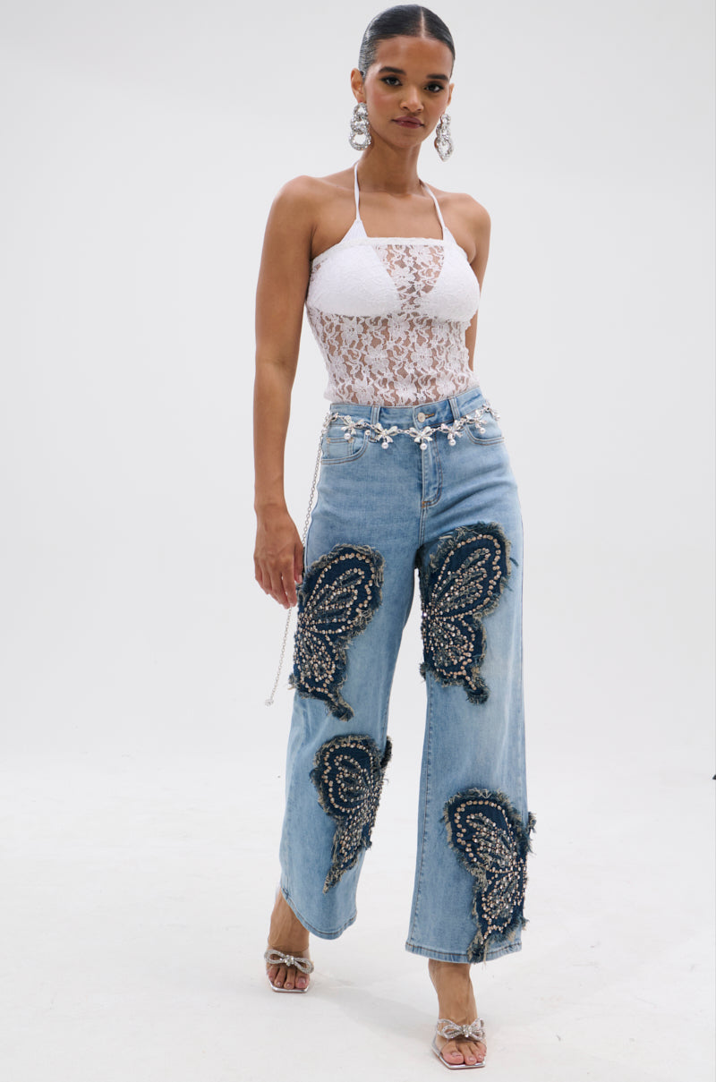A woman wears REBIRTH BUTTERFLY PATCH JEANS, light blue jeans with butterfly patches and beaded details, styled with a white lace halter top, large earrings, a jeweled belt, and heeled sandals. 5