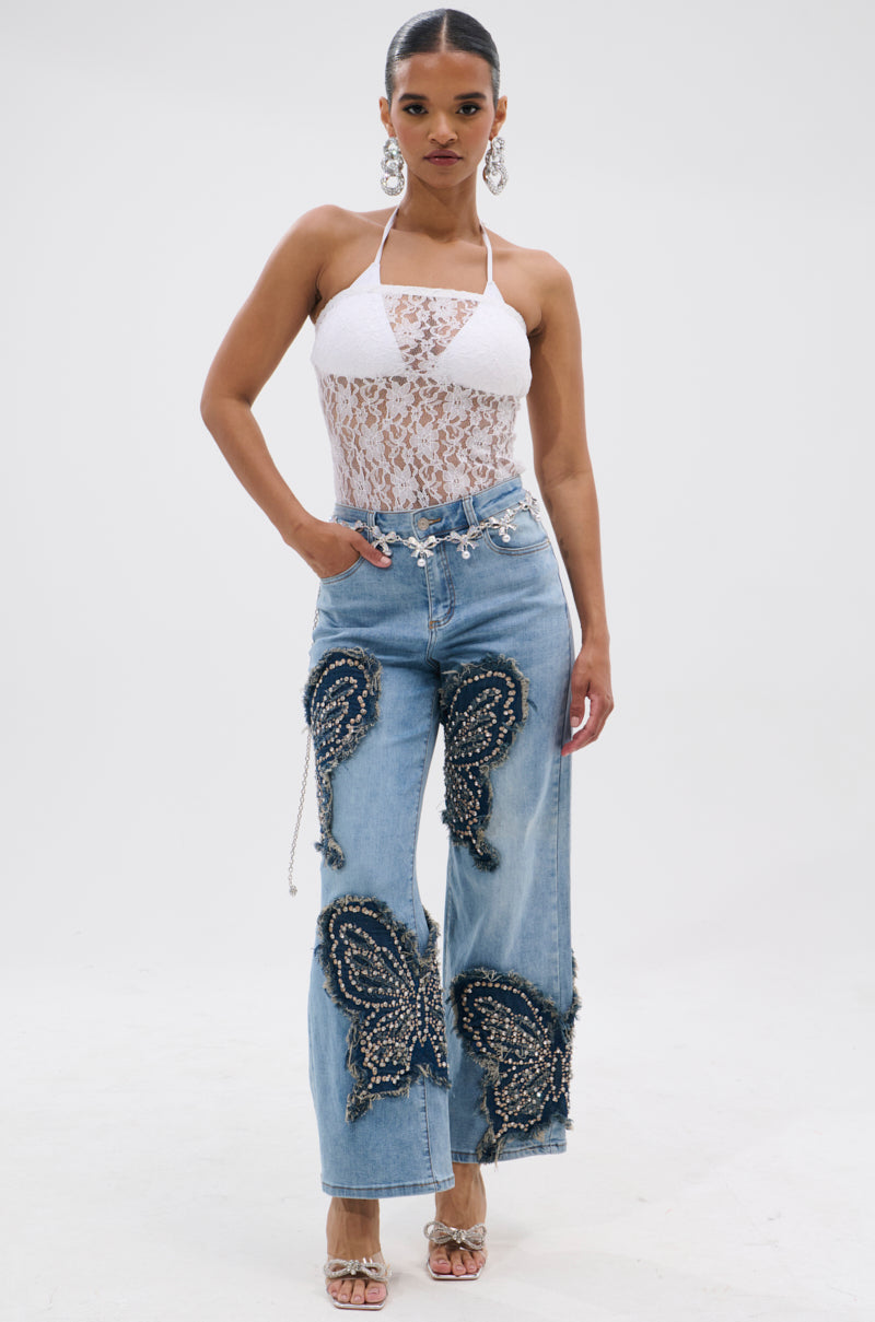 A woman models the REBIRTH BUTTERFLY PATCH JEANS, featuring light blue wide legs and butterfly patches, styled with a halter-neck white lace bodysuit, silver earrings, high heels, and her hair pulled back. 6