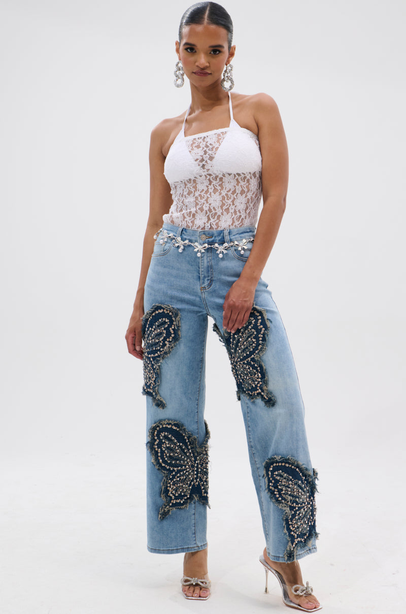 A woman poses in REBIRTH BUTTERFLY PATCH JEANS featuring black butterfly patches and pearls, paired with a white lace halter top, silver heels, and statement earrings, standing against a plain white background. 7