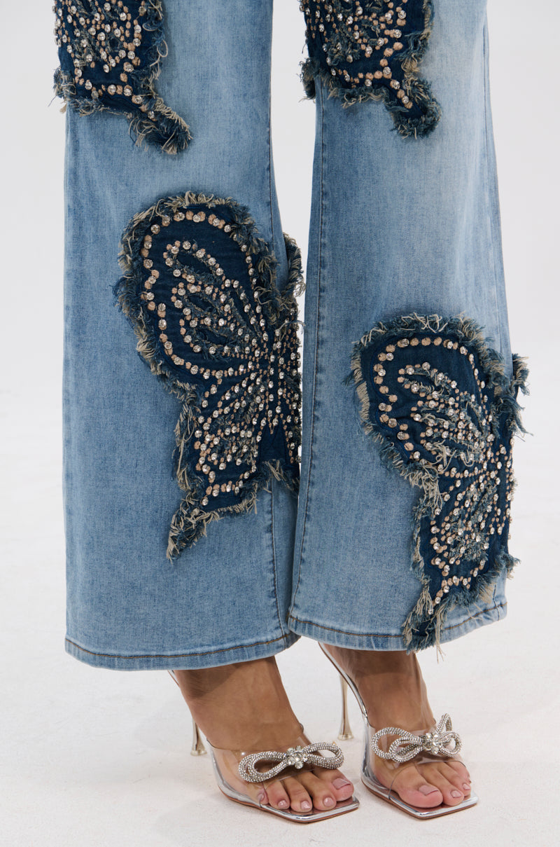 A close-up of someone wearing REBIRTH BUTTERFLY PATCH JEANS featuring large beaded butterfly patches and frayed denim, styled with clear high-heeled sandals decorated with bows. 8