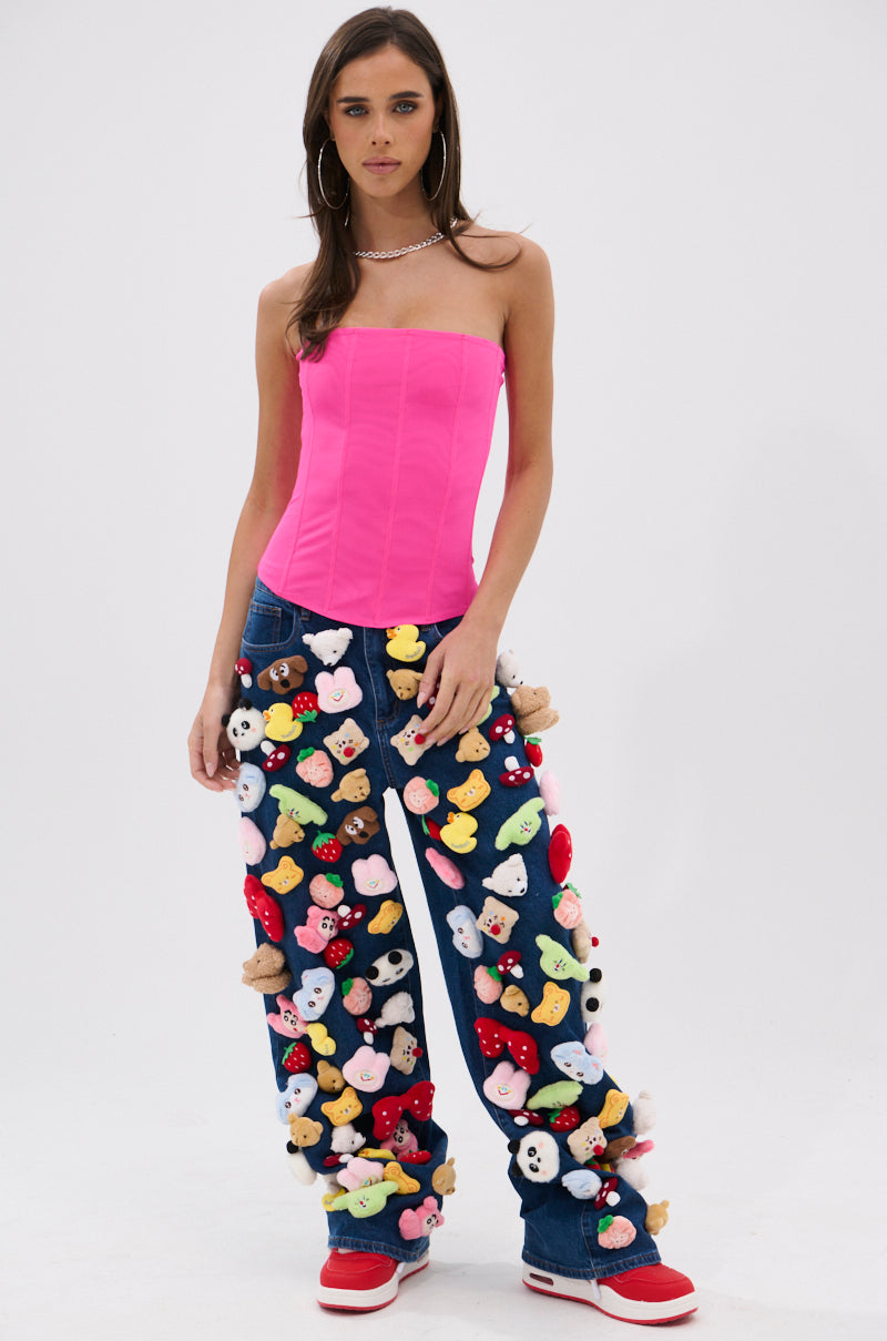 A woman with long brown hair wears a bright pink strapless top and the TEDDY BEAR DENIM PANT, featuring a wide-leg silhouette with colorful stuffed animal heads. She pairs them with red sneakers and a necklace, standing against a plain white background.