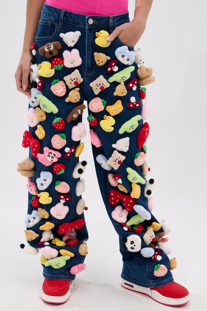Wearing the TEDDY BEAR DENIM PANT adorned with colorful plush toys, red and white sneakers, and a pink top, this person stands against a plain background, showing off a playful wide-leg silhouette.