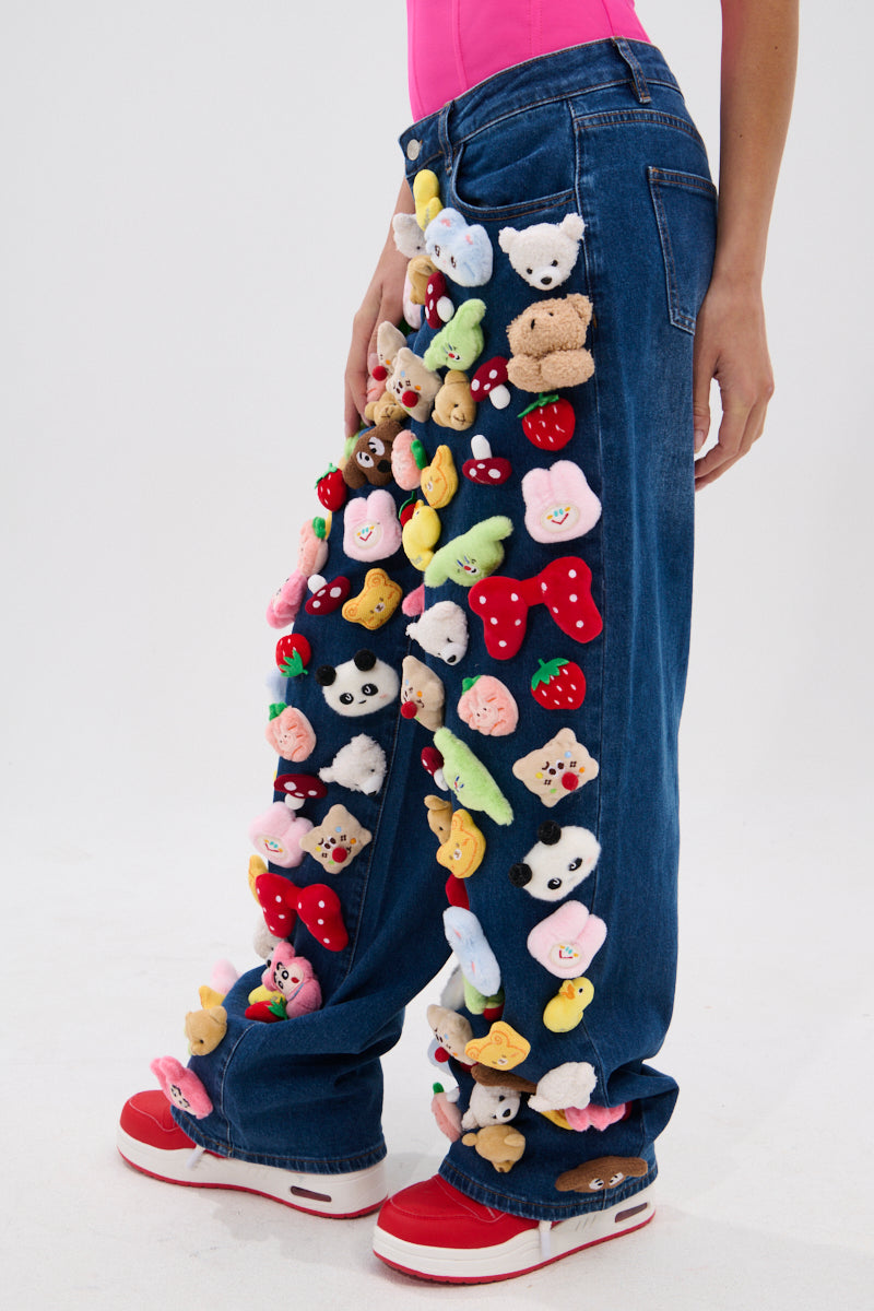 A person models the TEDDY BEAR DENIM PANT, high-rise jeans adorned with colorful plush toy animals and objects. They pair it with a pink top and red-and-white sneakers, with only their lower half visible against a plain backdrop.