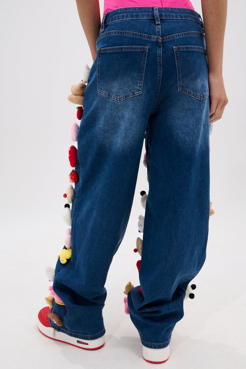 A person is seen from behind wearing the TEDDY BEAR DENIM PANT, featuring plush toys along the wide-leg side seams, styled with red and white sneakers.