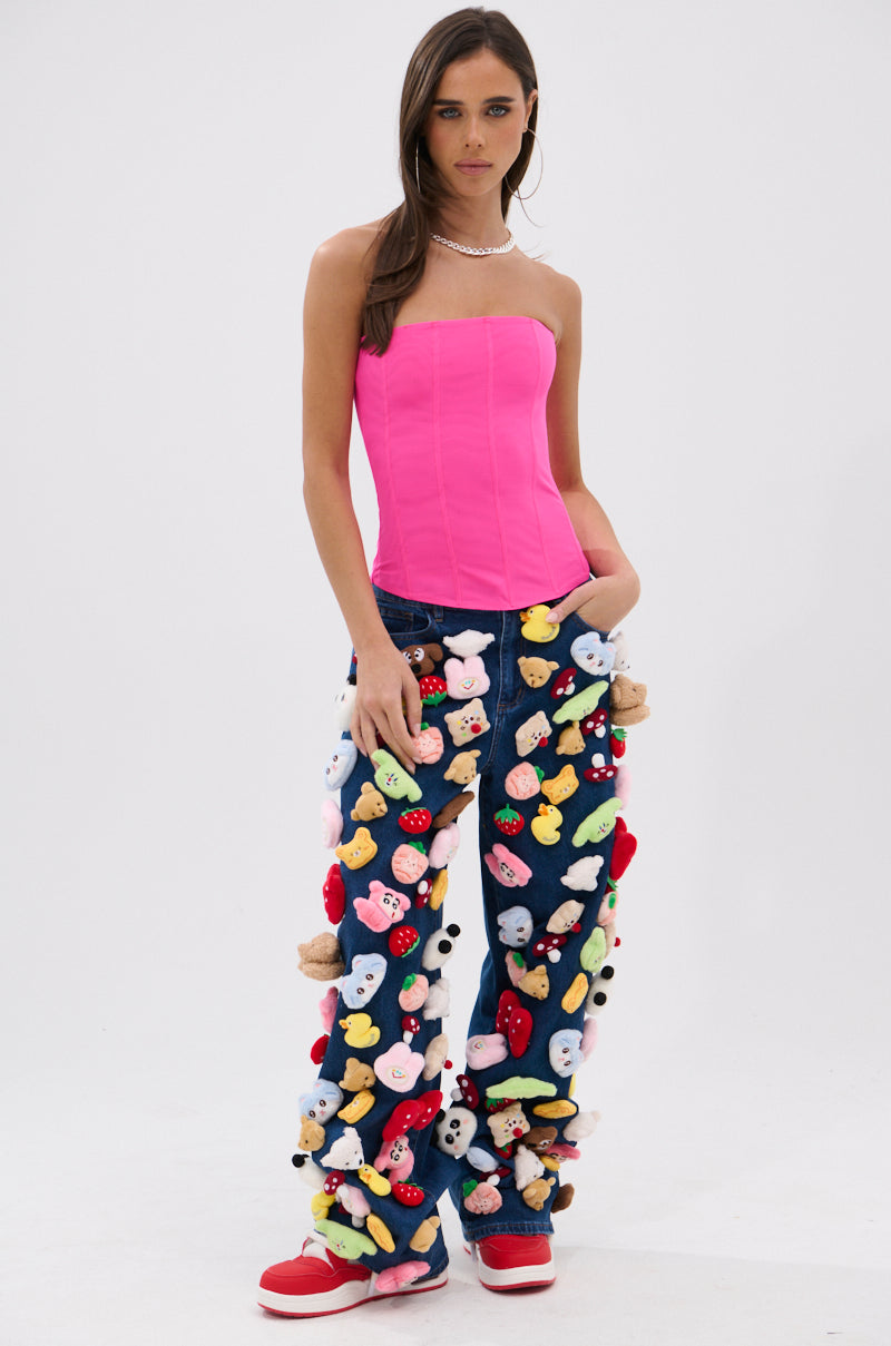 A woman in a bright pink strapless top and red sneakers wears the TEDDY BEAR DENIM PANT—high-rise, wide-leg jeans embellished with colorful stuffed animal toys—paired with a pearl necklace, against a plain white background.