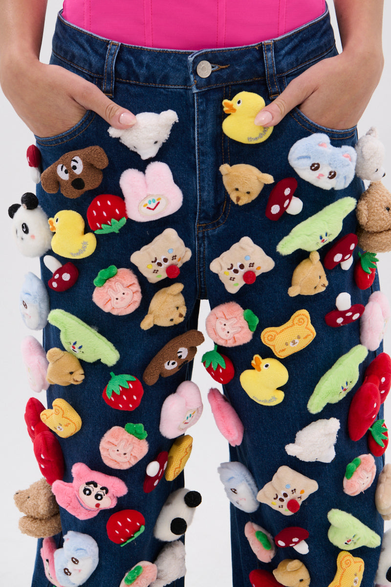 A person wearing the TEDDY BEAR DENIM PANT, decorated with colorful plush animals, fruits, and cartoon faces, stands with hands in pockets and pairs it with a bright pink top.