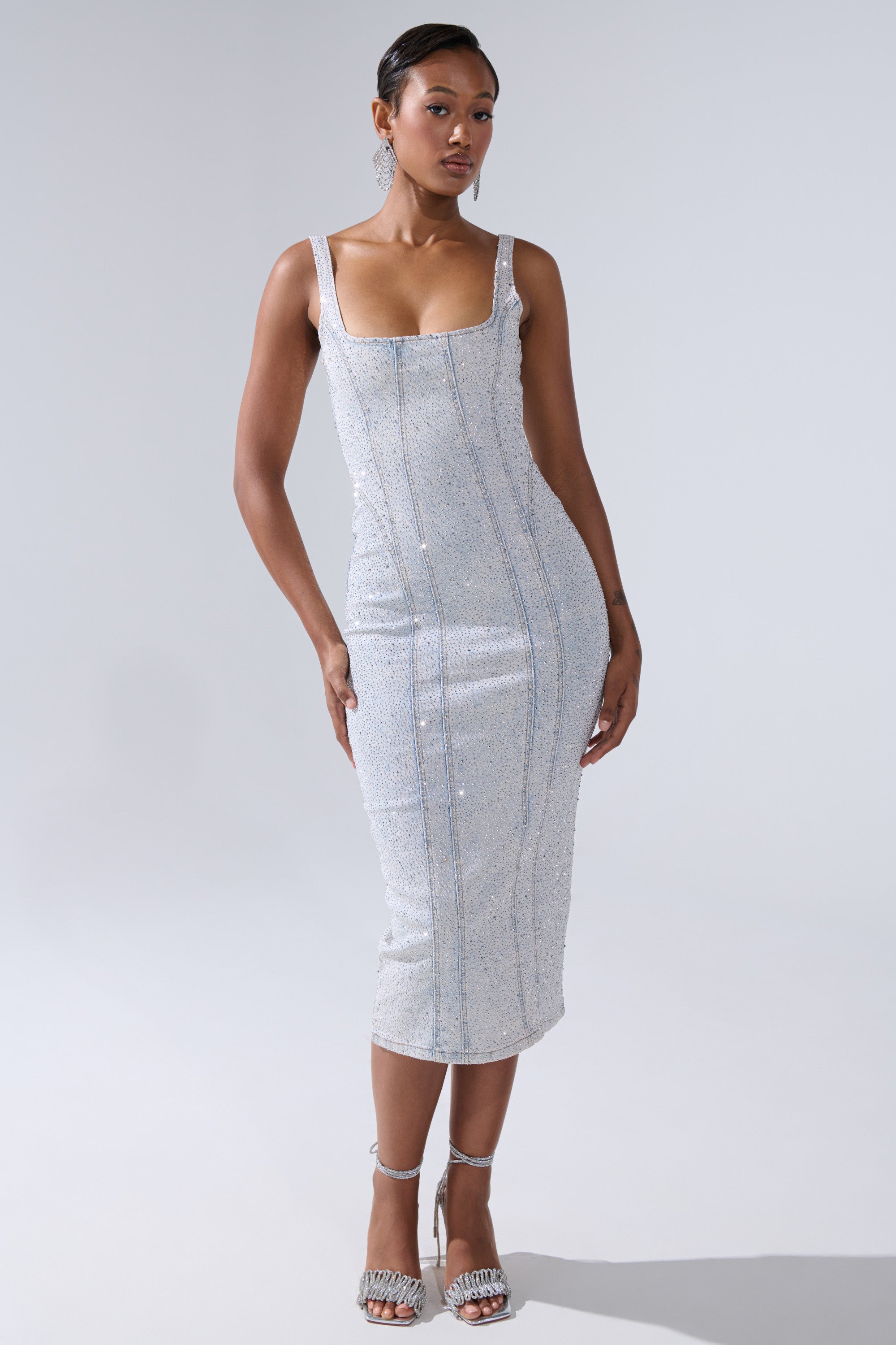 A woman poses against a plain background in the AFTER HOURS DENIM RHINESTONE MIDI DRESS, a fitted, sleeveless silver sequin piece with a square neckline. She pairs the dress with matching high heels, her hair pulled back, and one hand by her side.