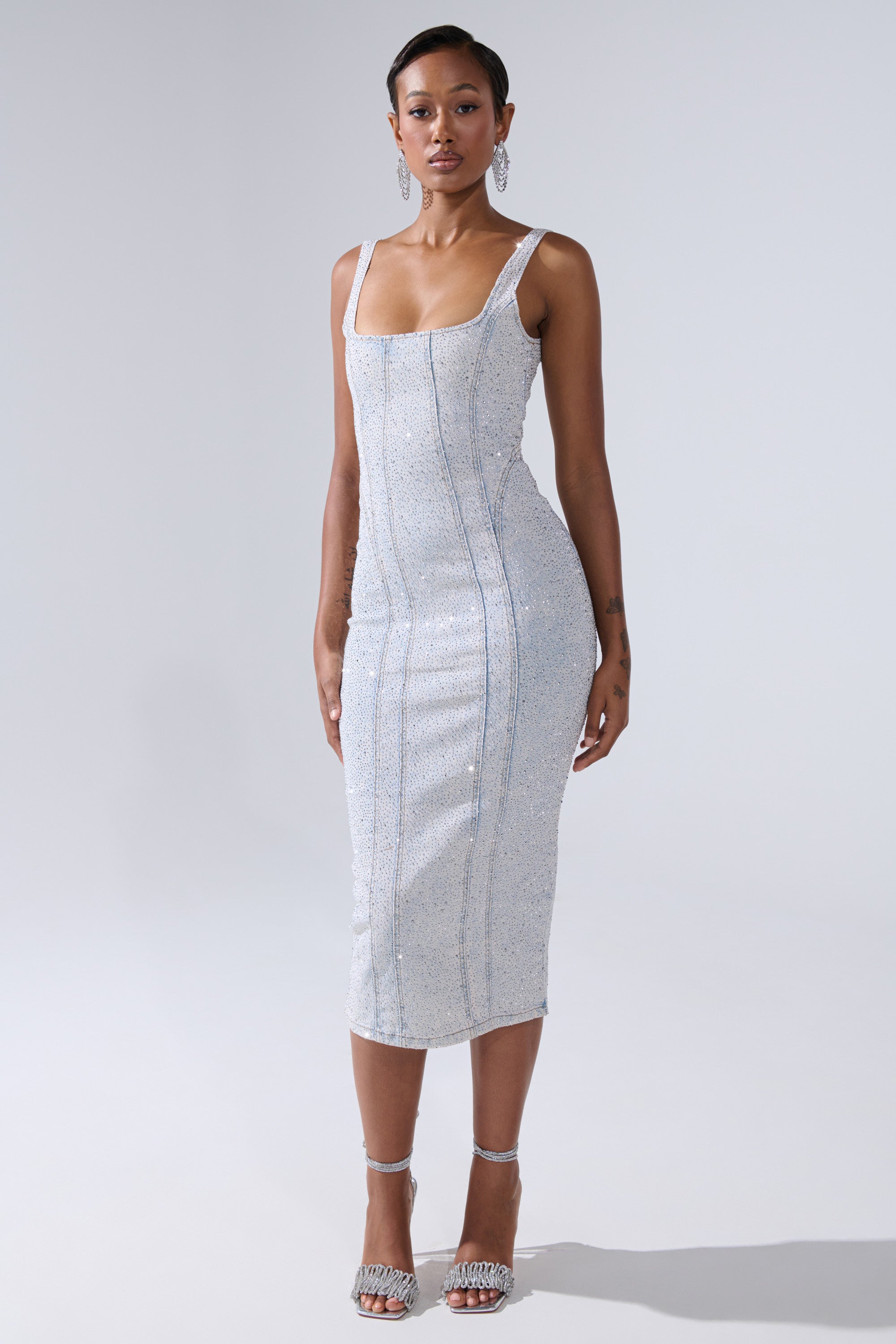 A woman stands against a plain background wearing the AFTER HOURS DENIM RHINESTONE MIDI DRESS with thin straps and rhinestone details. She pairs it with hoop earrings and embellished heels, making it a standout party statement.