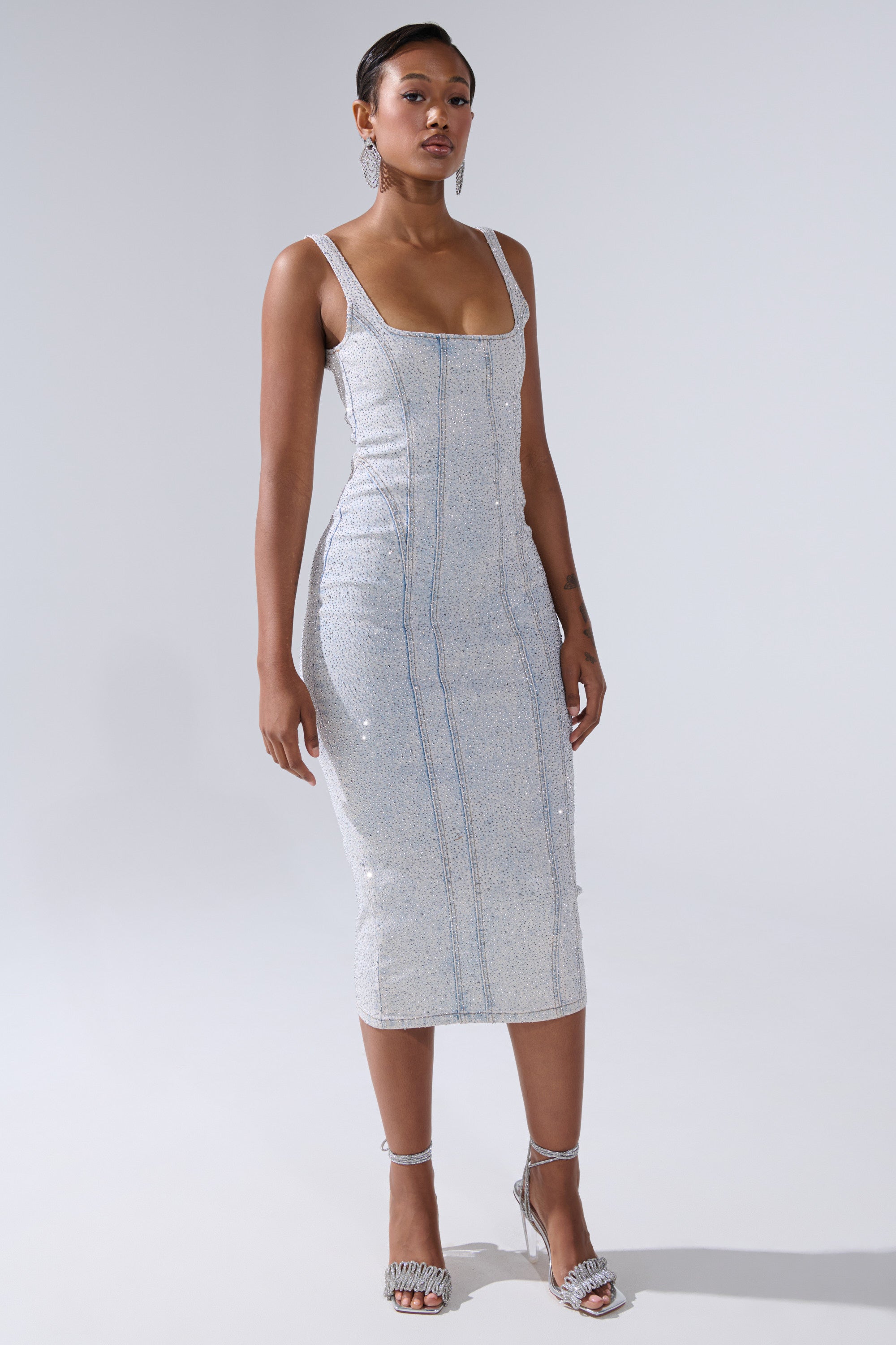 A woman poses against a plain light background in the AFTER HOURS DENIM RHINESTONE MIDI DRESS, a sparkly, fitted bodycon with thin straps and square neckline. She completes the look with matching high-heeled sandals and earrings.