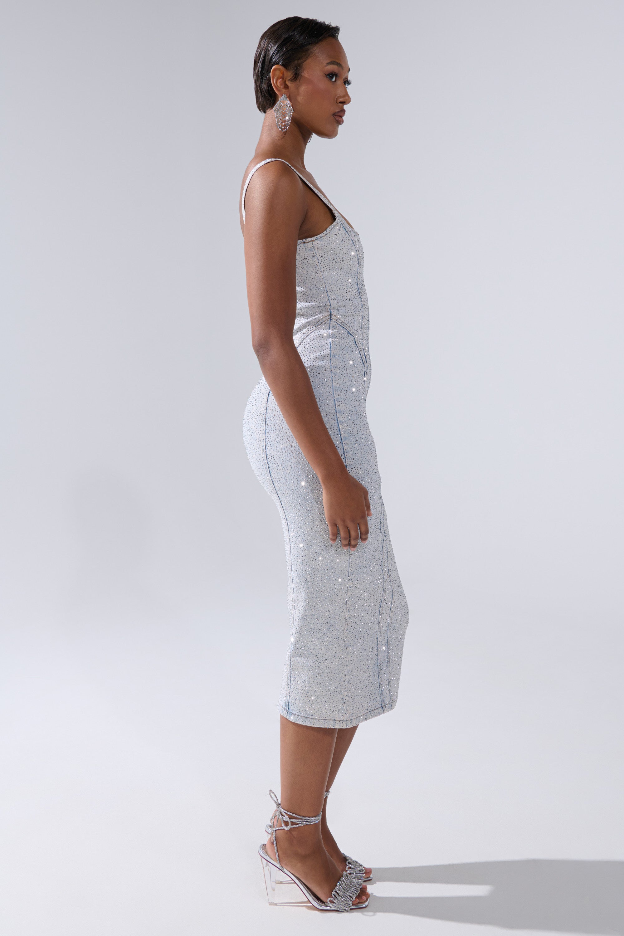 A woman in profile wears the AFTER HOURS DENIM RHINESTONE MIDI DRESS, a sparkly silver bodycon with thin straps. She has sleek short hair, hoop earrings, and silver ankle-strap heels against a plain light background.