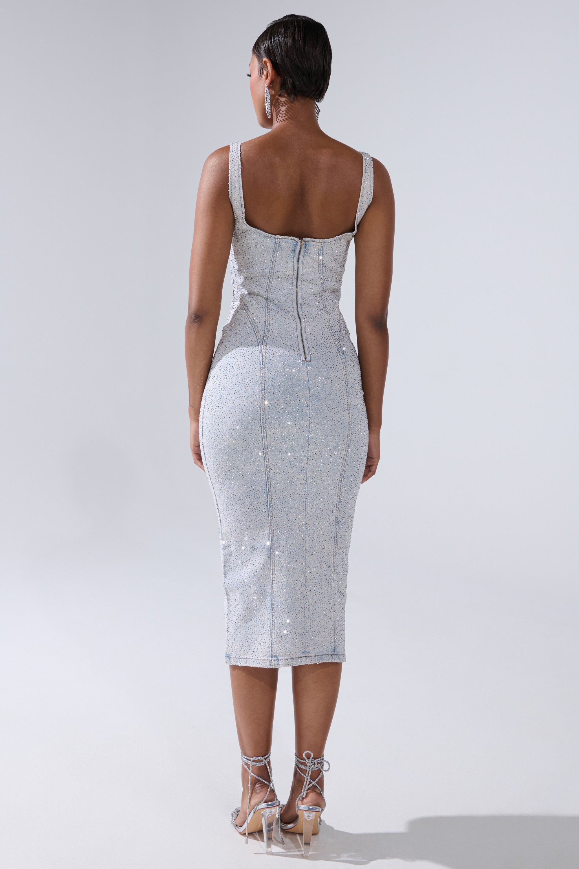 A woman with short hair faces away, wearing the AFTER HOURS DENIM RHINESTONE MIDI DRESS with a back zipper, styled with strappy high-heeled sandals and large hoop earrings against a plain background.